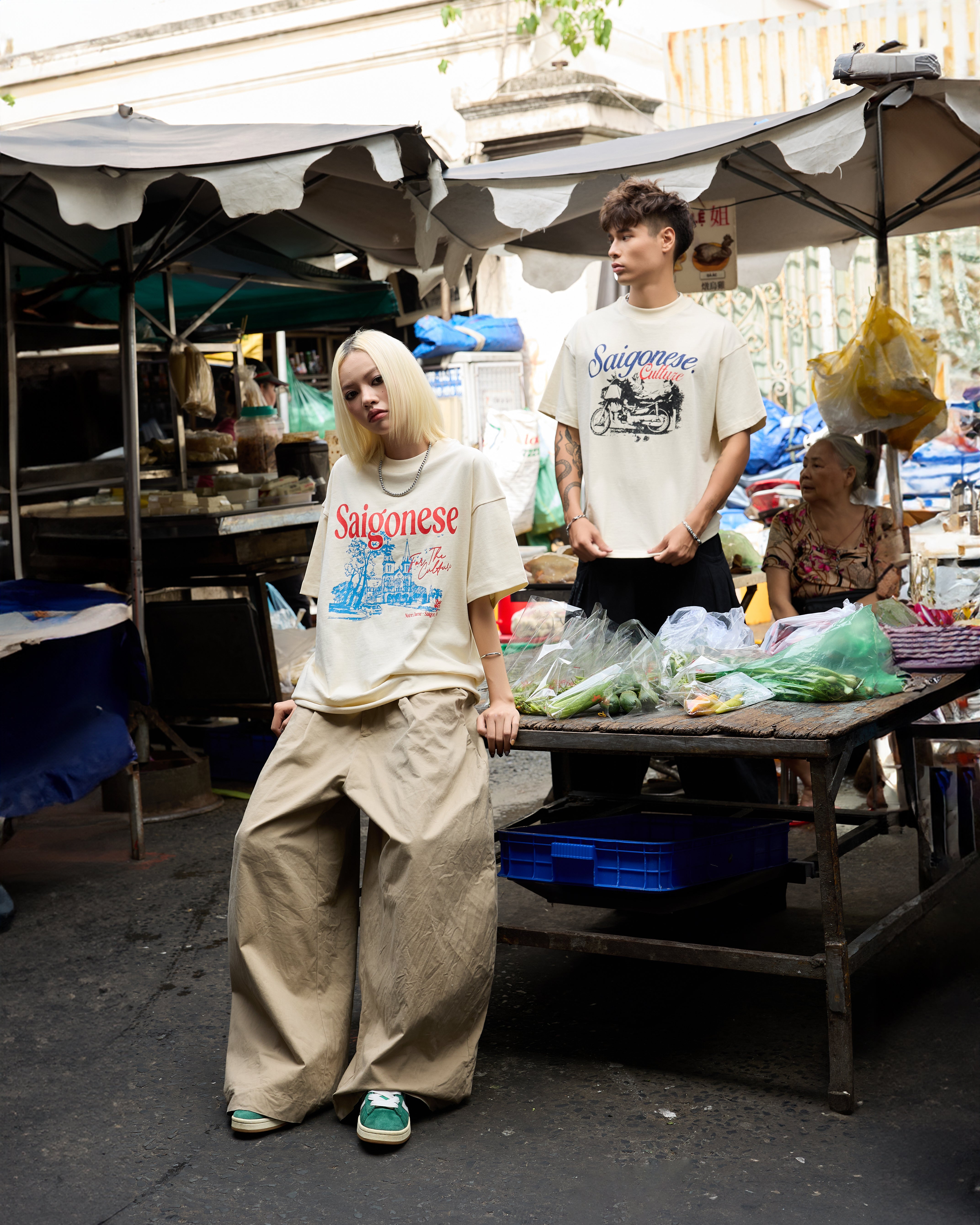 Two people wearing 'Saigonese' t-shirts in a market setting.