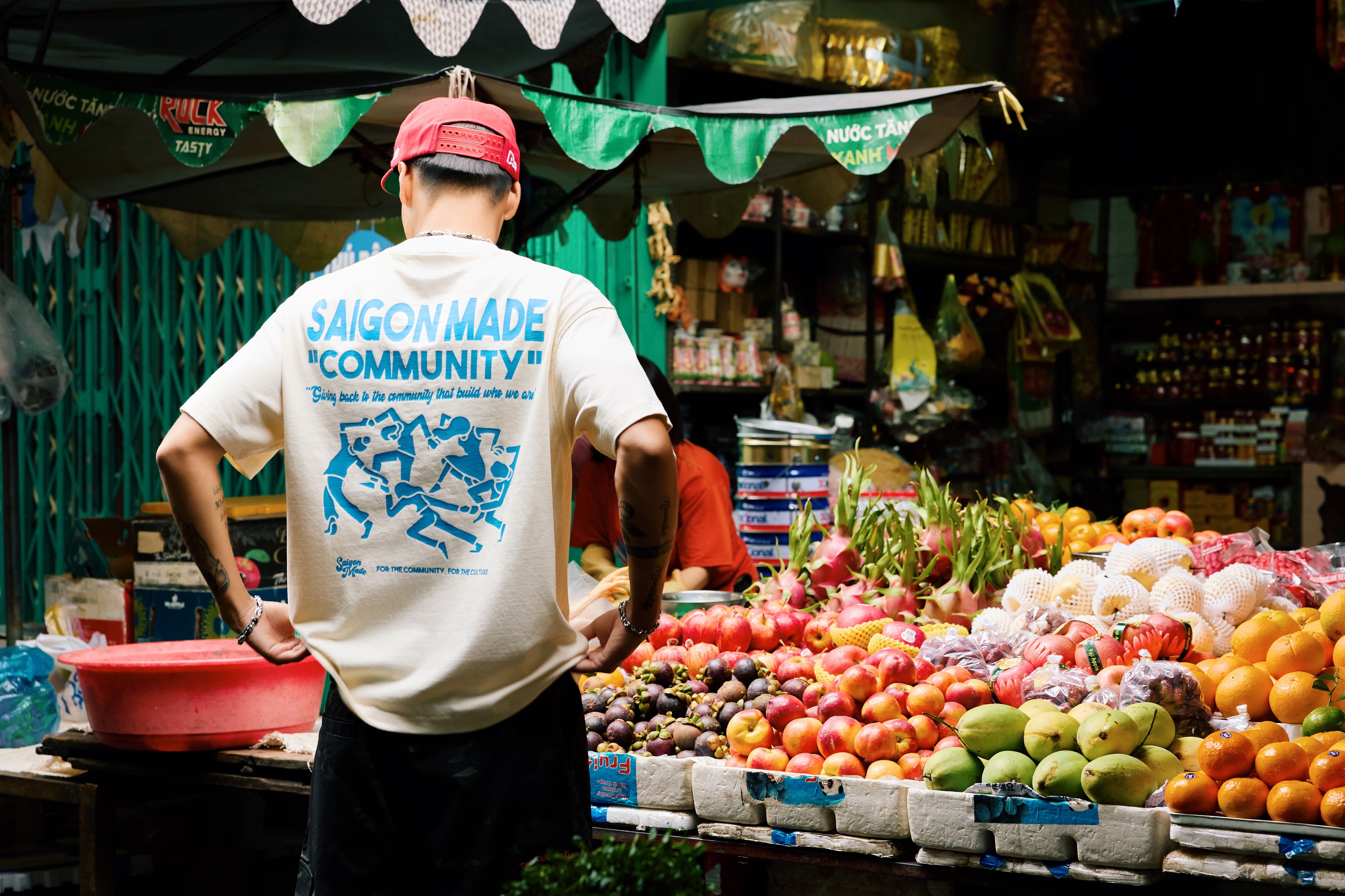 Person wearing a 'Saigon Made Community' shirt at a fruit stand.
