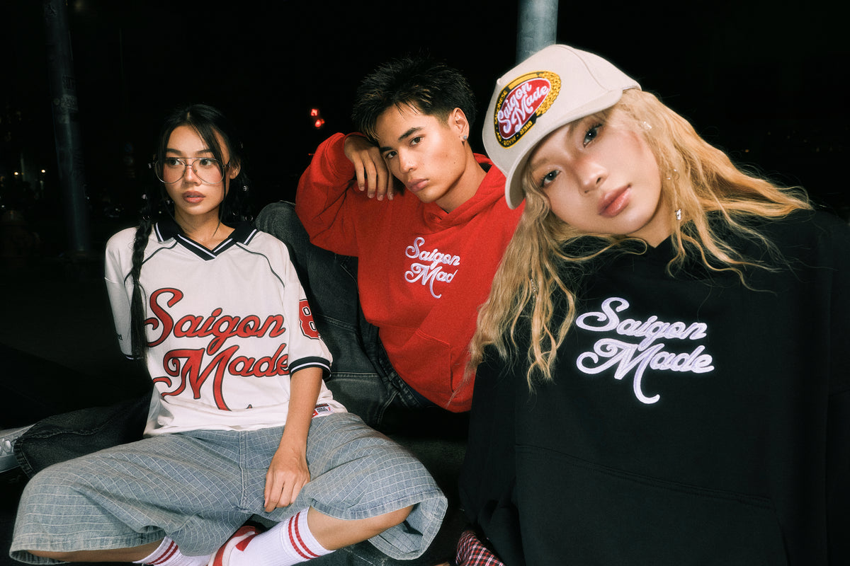 Three young models posing together at night wearing Saloon Made brand apparel, including a white baseball-style shirt with red script, a red hoodie, and a black sweatshirt with white script, set against a dark outdoor background.