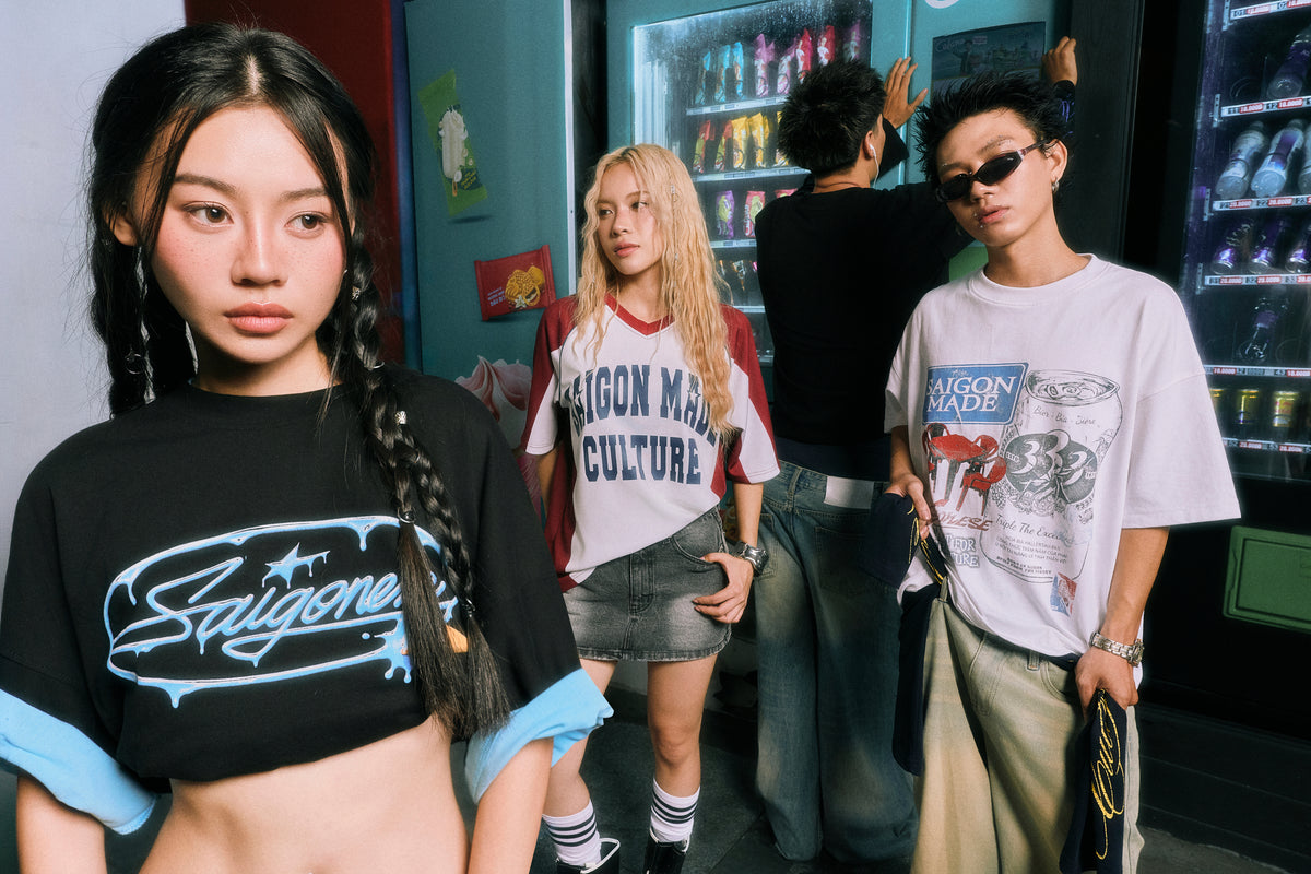 Three young women posing in streetwear T-shirts featuring the 'Saigon Made Culture' brand logo, set against a background with vending machines and a man.